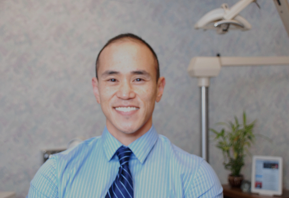 Dr. Hilehito Gene Lin smiling in his dental office, wearing a blue striped shirt and tie.